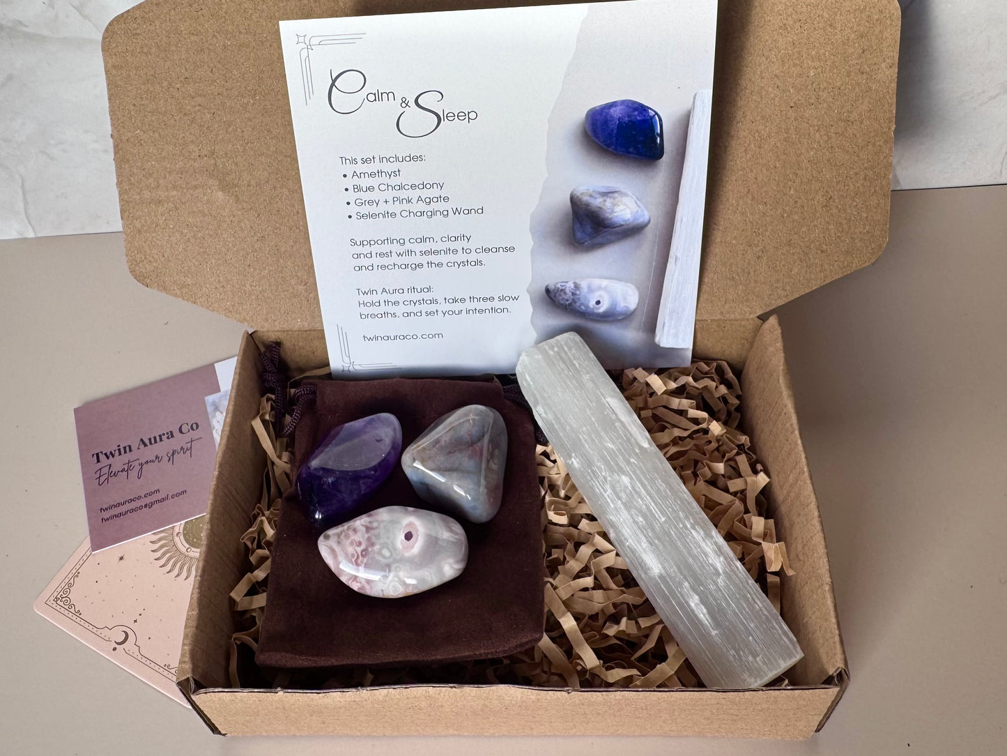 Box containing stones, a crystal, and a card with 'Calm & Sleep' branding on a neutral background. Amethyst, Blue Chalcedony, Grey and Pink agate, with Selenite wand and brown velvet bag. 