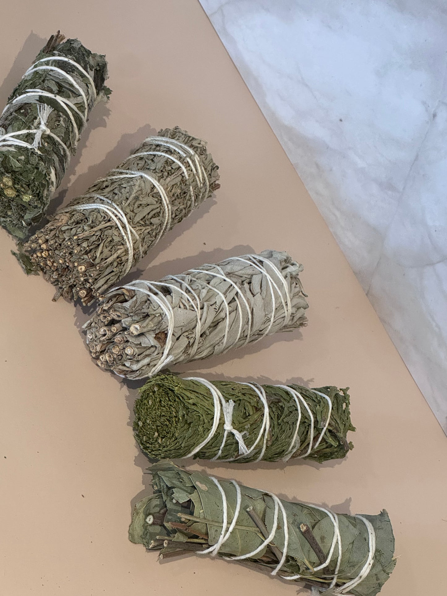 Five bundles of herbs, blue sage, white sage, eucalyptus, Mugwort, cedar, tied with white string on a beige surface with a marble background 