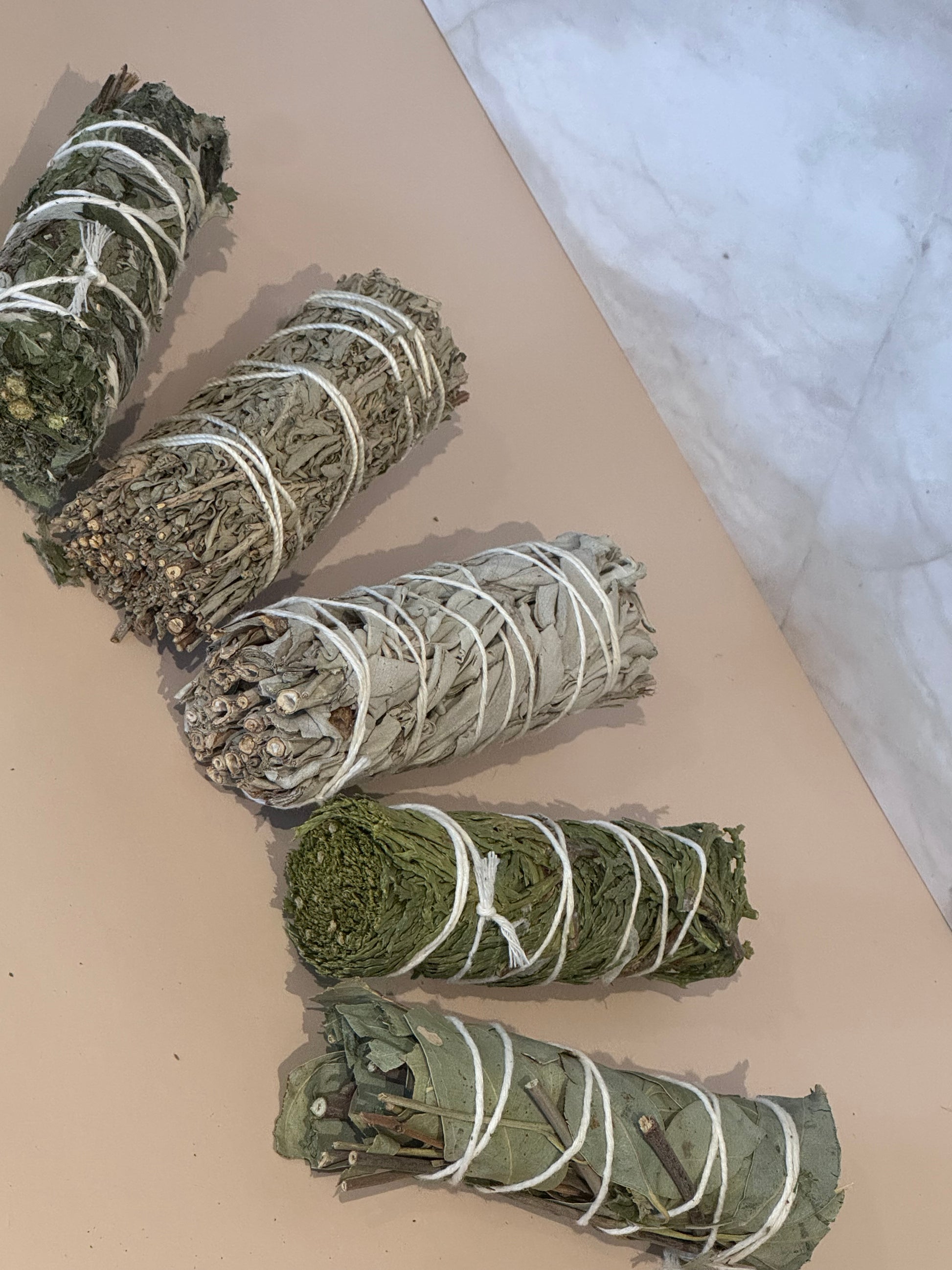 Five bundles of herbs, blue sage, white sage, eucalyptus, Mugwort, cedar, tied with white string on a beige surface with a marble background 
