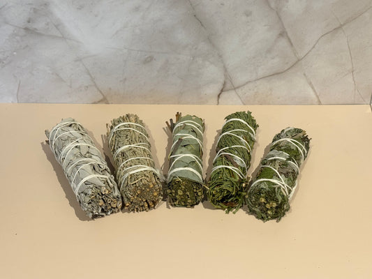 Five bundles of herbs, blue sage, white sage, eucalyptus, Mugwort, cedar, tied with white string on a beige surface with a marble background 